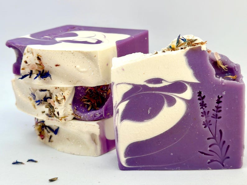 K&ouml;nnte beinhalten: Ein Stapel handgemachter Lavendelseifen mit einem Wirbelmuster in Violett- und Wei&szlig;t&ouml;nen. Die Seifen sind mit getrockneten Lavendelbl&uuml;ten verziert und haben ein Lavendelpflanzenmuster, das auf der Unterseite eingepr&auml;gt ist.