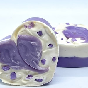 K&ouml;nnte beinhalten: Zwei herzf&ouml;rmige Seifen mit einem Wirbelmuster in Violett- und Wei&szlig;t&ouml;nen. Die Seifen haben kleine violette Flocken &uuml;berall.
