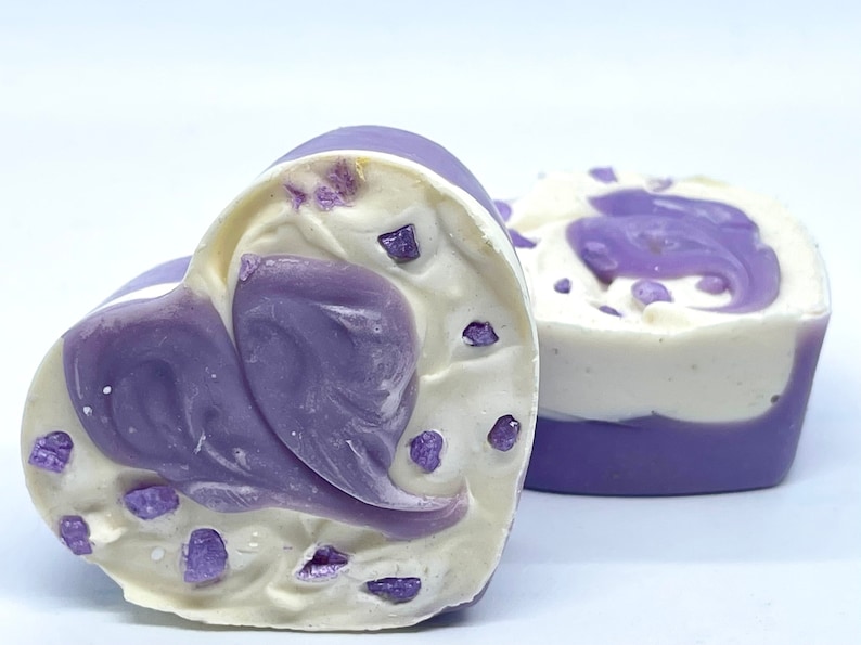 K&ouml;nnte beinhalten: Zwei herzf&ouml;rmige Seifen mit einem Wirbelmuster in Violett- und Wei&szlig;t&ouml;nen. Die Seifen haben kleine violette Flocken &uuml;berall.
