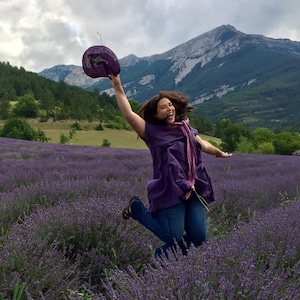 Pu&ograve; includere: Una donna con una camicia viola e jeans blu salta in un campo di fiori di lavanda. Tiene un cappello viola nella mano destra. Le montagne sono sullo sfondo.