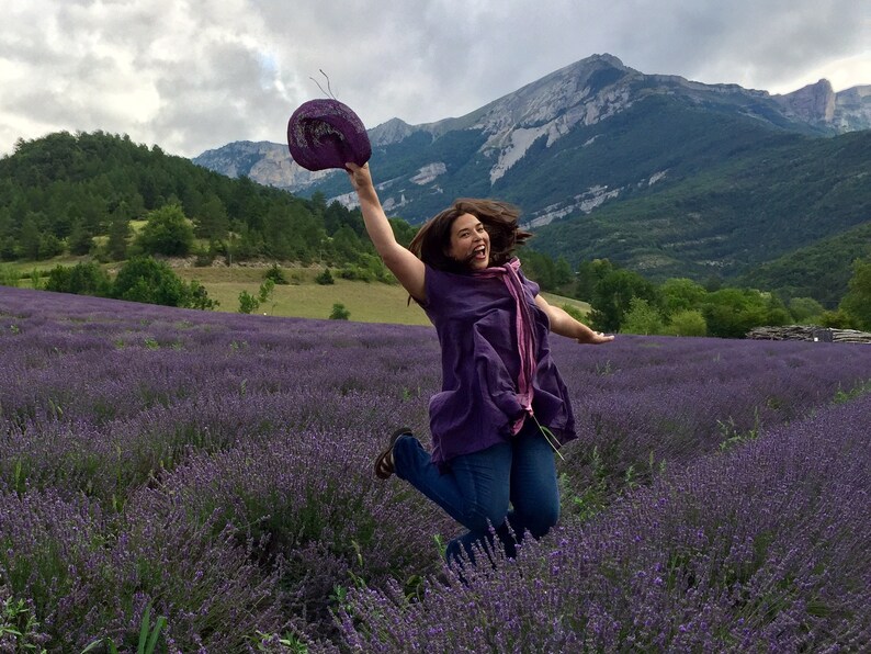 Pu&ograve; includere: Una donna con una camicia viola e jeans blu salta in un campo di fiori di lavanda. Tiene un cappello viola nella mano destra. Le montagne sono sullo sfondo.