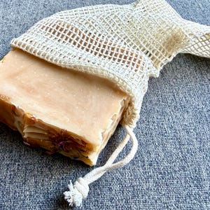 May include: A rectangular bar of soap with a light pink hue, partially encased in a cream-colored, open-weave soap saver bag. The soap has visible layers and embedded floral elements. A drawstring closure secures the bag.