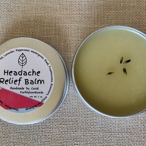 May include: A small, silver tin with a green label that reads "Headache Relief Balm" and lists ingredients including lavender, peppermint, wintergreen, basil, frankincense, rosemary, and orange. The tin is open and shows a green balm with dried lavender buds.
