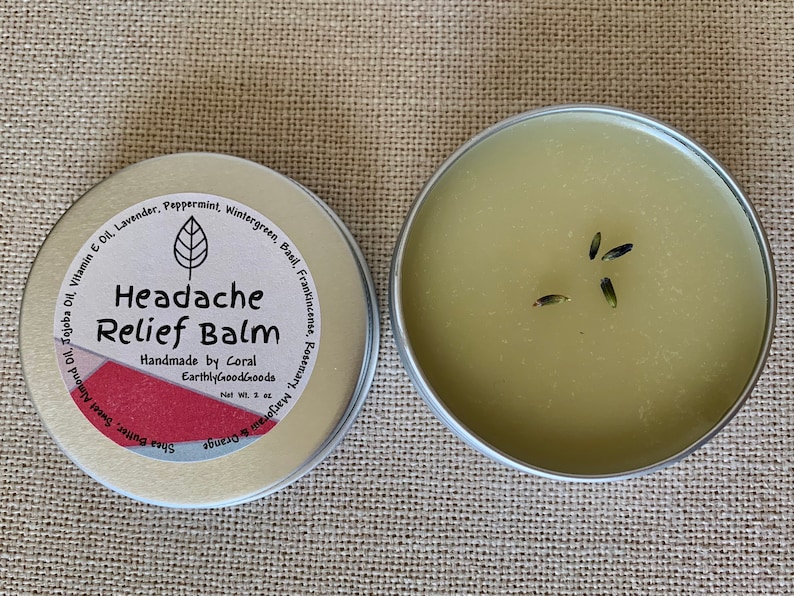 May include: A small, silver tin with a green label that reads "Headache Relief Balm" and lists ingredients including lavender, peppermint, wintergreen, basil, frankincense, rosemary, and orange. The tin is open and shows a green balm with dried lavender buds.