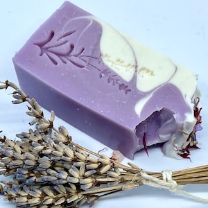 K&ouml;nnte beinhalten: Eine Seife mit Lavendel-Duft in Form eines Blocks mit einem Wirbelmuster in Violett- und Wei&szlig;t&ouml;nen. Die Seife hat ein gepr&auml;gtes Lavendelbl&uuml;tenmuster auf der Oberseite. Im Vordergrund befindet sich ein Bund getrockneter Lavendel.