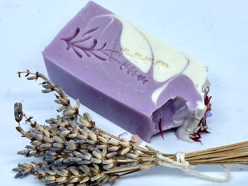 K&ouml;nnte beinhalten: Eine Seife mit Lavendel-Duft in Form eines Blocks mit einem Wirbelmuster in Violett- und Wei&szlig;t&ouml;nen. Die Seife hat ein gepr&auml;gtes Lavendelbl&uuml;tenmuster auf der Oberseite. Im Vordergrund befindet sich ein Bund getrockneter Lavendel.