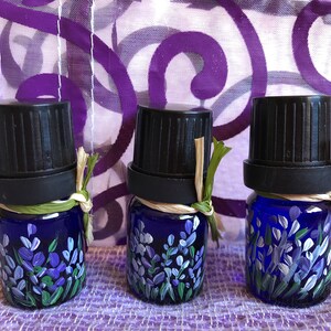 Pu&ograve; includere: Tre piccoli flaconi di vetro blu con tappi neri e disegni di lavanda dipinti a mano. I flaconi sono legati con spago e hanno un aspetto rustico e naturale.