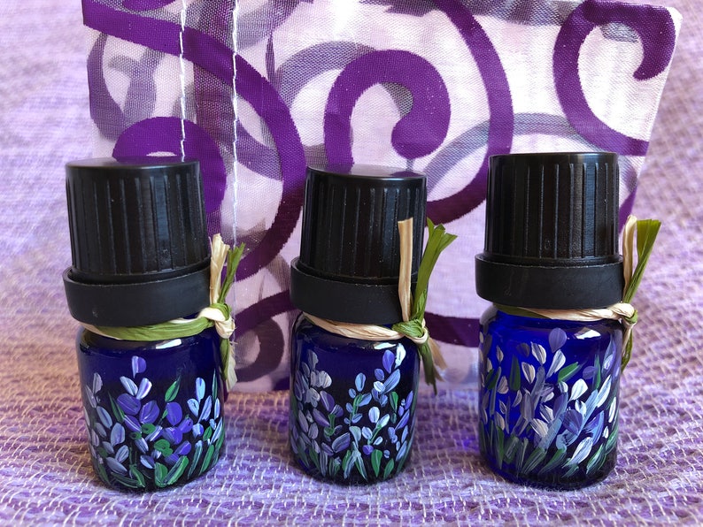 Pu&ograve; includere: Tre piccoli flaconi di vetro blu con tappi neri e disegni di lavanda dipinti a mano. I flaconi sono legati con spago e hanno un aspetto rustico e naturale.