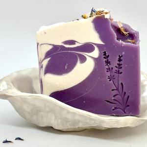 K&ouml;nnte beinhalten: Eine violette und wei&szlig;e Marmor-Seifenstange mit einem Lavendel-Design, die in einer wei&szlig;en Keramik-Seifenschale liegt.