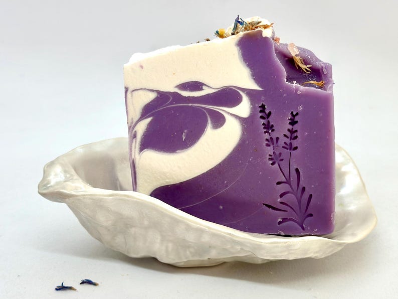 K&ouml;nnte beinhalten: Eine violette und wei&szlig;e Marmor-Seifenstange mit einem Lavendel-Design, die in einer wei&szlig;en Keramik-Seifenschale liegt.