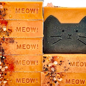 May include: A stack of six orange-colored soap bars with the word "MEOW!" embossed on each bar. Next to the stack is a single orange-colored soap bar with a black cat face design.