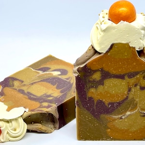 May include: Two handmade soap bars with a swirl pattern of brown, orange, and purple. The soap bars are topped with a white whipped cream-like topping and an orange sphere.
