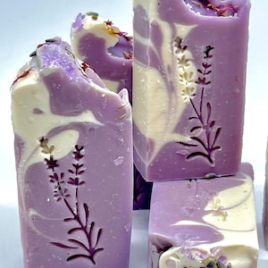 K&ouml;nnte beinhalten: Vier handgemachte Lavendelseifen mit einem Swirl-Design in Violett- und Wei&szlig;t&ouml;nen. Jede Seife hat ein gepr&auml;gtes Lavendelbl&uuml;tenmuster auf der Vorderseite. Die Seife ist mit getrockneten Lavendelbl&uuml;ten und rosa Himalaya-Salz verziert.