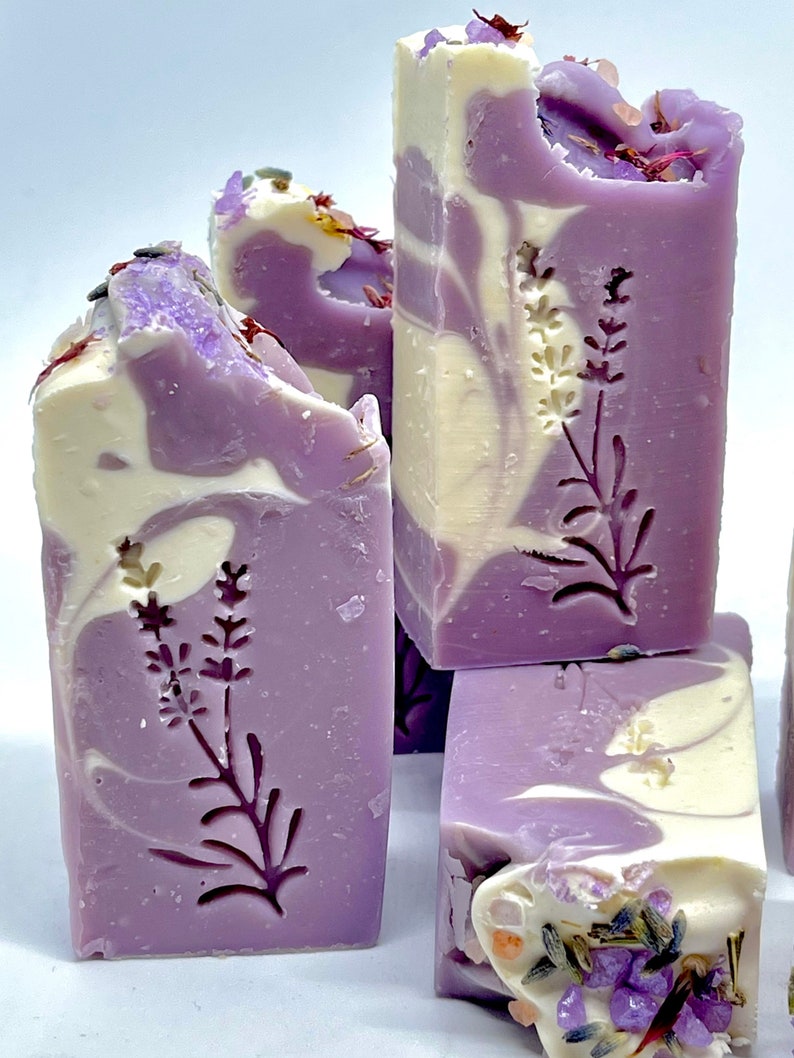 K&ouml;nnte beinhalten: Vier handgemachte Lavendelseifen mit einem Swirl-Design in Violett- und Wei&szlig;t&ouml;nen. Jede Seife hat ein gepr&auml;gtes Lavendelbl&uuml;tenmuster auf der Vorderseite. Die Seife ist mit getrockneten Lavendelbl&uuml;ten und rosa Himalaya-Salz verziert.