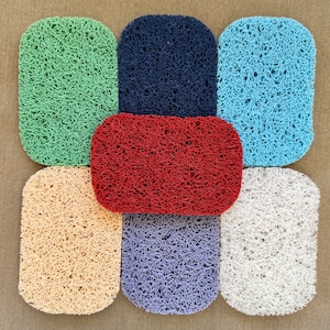 May include: A collection of seven rectangular, textured soap savers in various colors: green, navy blue, light blue, red, beige, purple, and white. The soap savers are arranged on a neutral-toned surface.