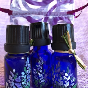 Pu&ograve; includere: Tre piccoli flaconi di vetro blu con tappi neri, ognuno dipinto a mano con fiori di lavanda. I flaconi sono su una superficie di tessuto viola.