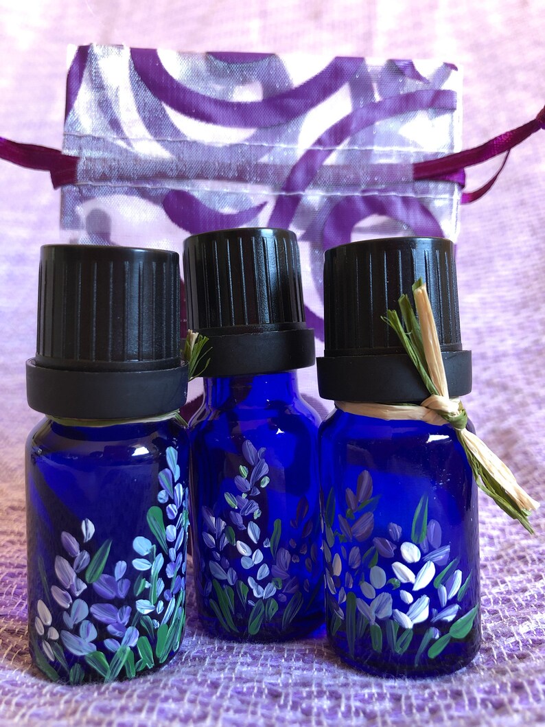 Pu&ograve; includere: Tre piccoli flaconi di vetro blu con tappi neri, ognuno dipinto a mano con fiori di lavanda. I flaconi sono su una superficie di tessuto viola.
