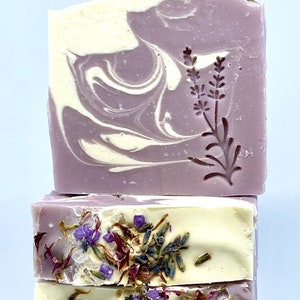 K&ouml;nnte beinhalten: Drei St&uuml;ck Lavendelseife mit violetten und wei&szlig;en Wirbeln und getrockneten Lavendelbl&uuml;ten. Die obere Seife hat ein Lavendelbl&uuml;tenmuster.