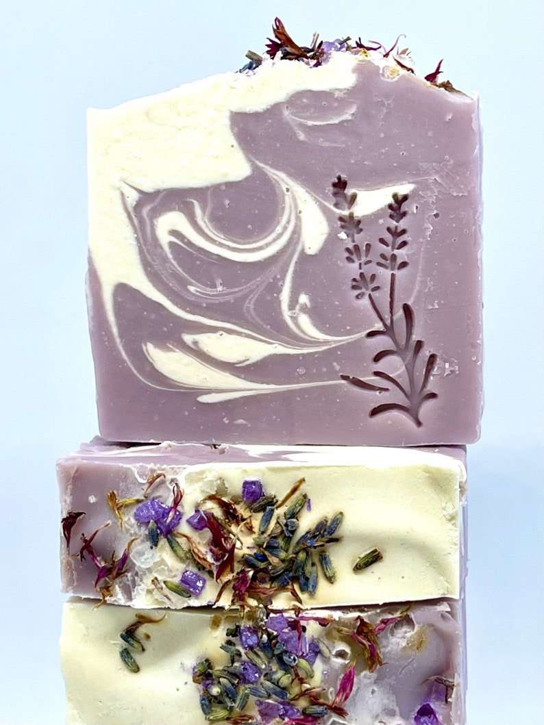 K&ouml;nnte beinhalten: Drei St&uuml;ck Lavendelseife mit violetten und wei&szlig;en Wirbeln und getrockneten Lavendelbl&uuml;ten. Die obere Seife hat ein Lavendelbl&uuml;tenmuster.