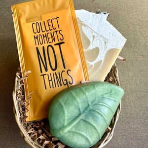 May include: A wicker basket filled with brown shredded paper, a green leaf-shaped soap, a white and brown swirl soap, and a yellow pouch with the text "Collect Moments Not Things" printed on it.