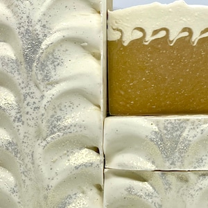 May include: Four bars of handmade soap. Two bars are white with silver glitter and a swirl design. The other two bars are a light brown color with a white swirl design.