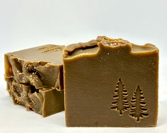 May include: Two bars of brown soap with gold glitter. The soap has a design of two pine trees on one bar.