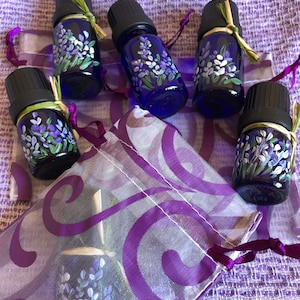 Pu&ograve; includere: Cinque piccoli flaconi di vetro blu con tappi neri e disegni di fiori di lavanda sono disposti su un tessuto a motivi viola e bianchi. I flaconi sono legati con uno spago e hanno un aspetto rustico e fatto a mano.