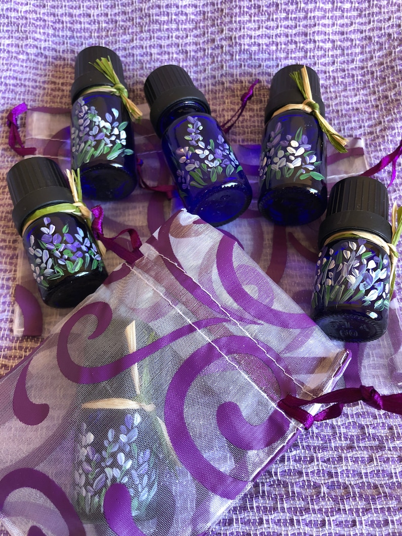 Pu&ograve; includere: Cinque piccoli flaconi di vetro blu con tappi neri e disegni di fiori di lavanda sono disposti su un tessuto a motivi viola e bianchi. I flaconi sono legati con uno spago e hanno un aspetto rustico e fatto a mano.
