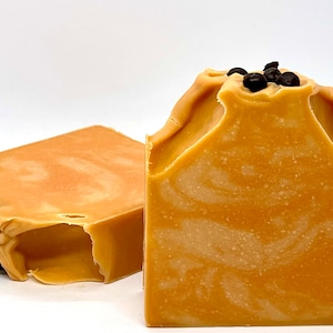 May include: Two bars of handmade soap, one with a swirl pattern and the other with a plain surface. Both bars are a light orange colour and have dark brown specks on top.