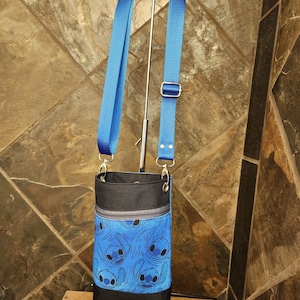 May include: A blue crossbody bag featuring a repeating pattern of a cartoon character. The bag has a black top and bottom, a zippered pocket, and a blue adjustable strap. The bag is hanging from a silver stand.