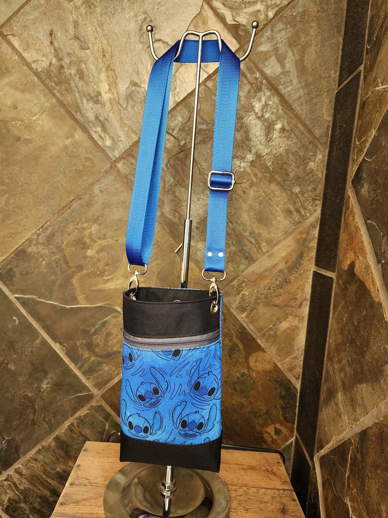May include: A blue crossbody bag featuring a repeating pattern of a cartoon character. The bag has a black top and bottom, a zippered pocket, and a blue adjustable strap. The bag is hanging from a silver stand.