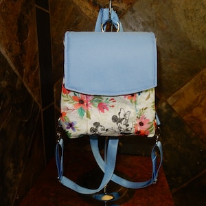 May include: A small, light blue backpack with a flap closure. The lower portion features a floral pattern with pink, orange, and purple flowers. Mickey and Minnie Mouse are depicted in black and white.