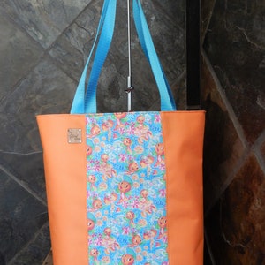 May include: Orange tote bag with light blue handles. The center panel features a colorful pattern of orange characters and flowers on a blue background. A small, rectangular tag is attached to the front.