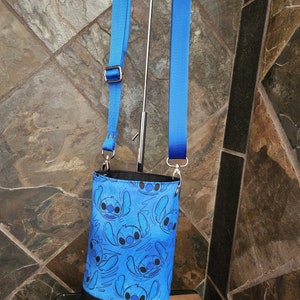 May include: A blue crossbody bag featuring a repeating pattern of a cartoon character. The bag has a black bottom and a blue adjustable strap. The bag is hanging on a metal stand.