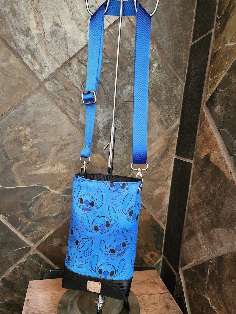 May include: A blue crossbody bag featuring a repeating pattern of a cartoon character. The bag has a black bottom and a blue adjustable strap. The bag is hanging on a metal stand.