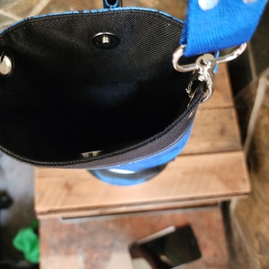 May include: A blue and black cylindrical crossbody bag with a blue strap. The bag has a black interior and a magnetic snap closure. The strap is attached with silver-colored metal hardware.