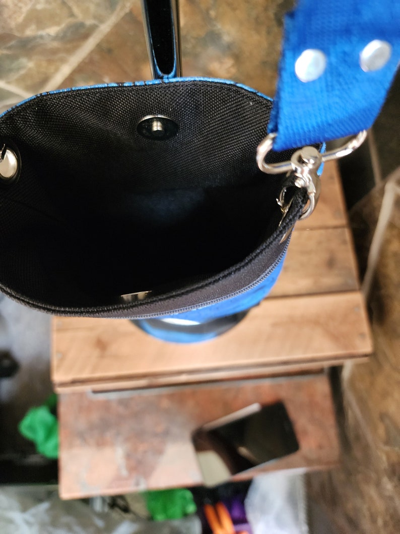May include: A blue and black cylindrical crossbody bag with a blue strap. The bag has a black interior and a magnetic snap closure. The strap is attached with silver-colored metal hardware.