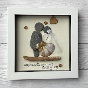 May include: Framed pebble art depicting a bride and groom, crafted from gray pebbles. The bride has a veil and holds a seashell. Wooden hearts and the text "Congratulations on your Wedding Day" are included in the artwork.