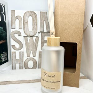 May include: A clear glass bottle with a wooden lid and a label that reads "La scent Lemongrass & Persian Lime Handmade Reed Diffuser". The bottle is sitting on a white surface with a brown box behind it.