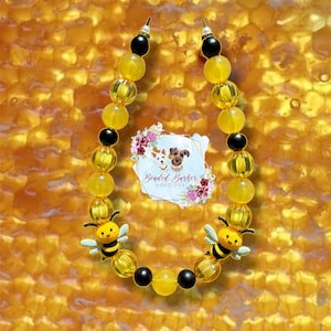 Peut inclure: Un collier pour animaux de compagnie à perles jaunes avec des accents noirs et deux breloques abeilles. Le collier porte le texte "Beaded Barker Boutique" sur une étiquette blanche avec des accents floraux. Le fond est un motif en nid d'abeilles.