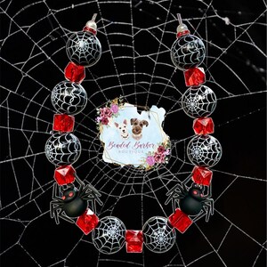 Puede incluir: Un collar temático de Halloween con cuentas de telaraña en blanco y negro, cuentas rojas en forma de cubo y abalorios de araña negros. El collar está colocado sobre un fondo negro con un diseño de telaraña blanca. El logotipo de Beaded Barker Boutique está en el centro.