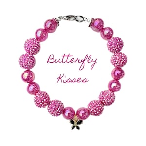 K&ouml;nnte beinhalten: Rosa Perlenarmband mit einem Schmetterlingsanh&auml;nger. Die Perlen sind irisierend und haben eine glitzernde Oberfl&auml;che. Das Armband hat einen silbernen Verschluss und der Text "Butterfly Kisses" steht in der Mitte.