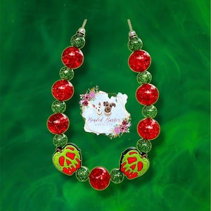 Op de afbeelding: Een halsband voor honden met rode en groene kralen. De halsband heeft twee groene appelbedels met een rood gifontwerp. De halsband is van Beaded Barker Boutique.