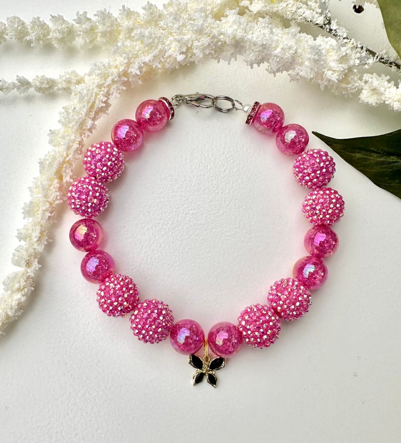 K&ouml;nnte beinhalten: Rosa- und regenbogenfarbenes Perlenarmband mit einem goldenen Schmetterlingsanh&auml;nger.