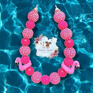May include: A beaded necklace featuring alternating hot pink rhinestone-encrusted spheres and smooth round beads. Two pink flamingo charms are positioned near the bottom, and a logo tag reading Beaded Barker Boutique is centered.