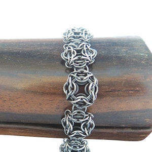 May include: A silver chainmail bracelet with a woven pattern. The bracelet is displayed on a brown wooden surface.