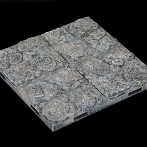 May include: A square tile made of gray stone with a textured surface. The tile is designed to look like a cobblestone path.