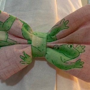 May include: A handmade bow tie with a pink fabric patterned with green frogs. The bow tie is tied in the centre with a matching fabric band. The bow tie is likely for a pet or child.