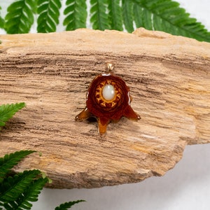 May include: A brown and orange resin pendant with a white opal stone in the center. The pendant is shaped like a stylized tree with a circular base and three branches. The pendant is sitting on a piece of driftwood.