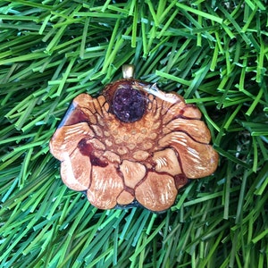 Puede incluir: Un colgante de madera marrón y blanco con un centro de resina púrpura. El colgante tiene forma de flor y tiene una anilla de oro.